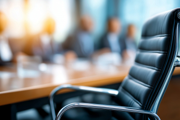 An empty leather chair beside a boardroom table, with a meeting in progress out of focus behind it.