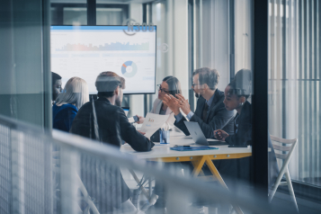 Business Leaders Sit Around a Conference Table Reviewing Charts on a Large Screen, Debating Results, Setting Priorities, and Planning Next Steps While Using Laptops, Printed Reports, and Notes During