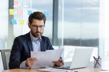 Serious thinking businessman behind paper work, mature experienced boss working with reports and financial documents, financier inside office with laptop, reading contract, thoughtfully checking.